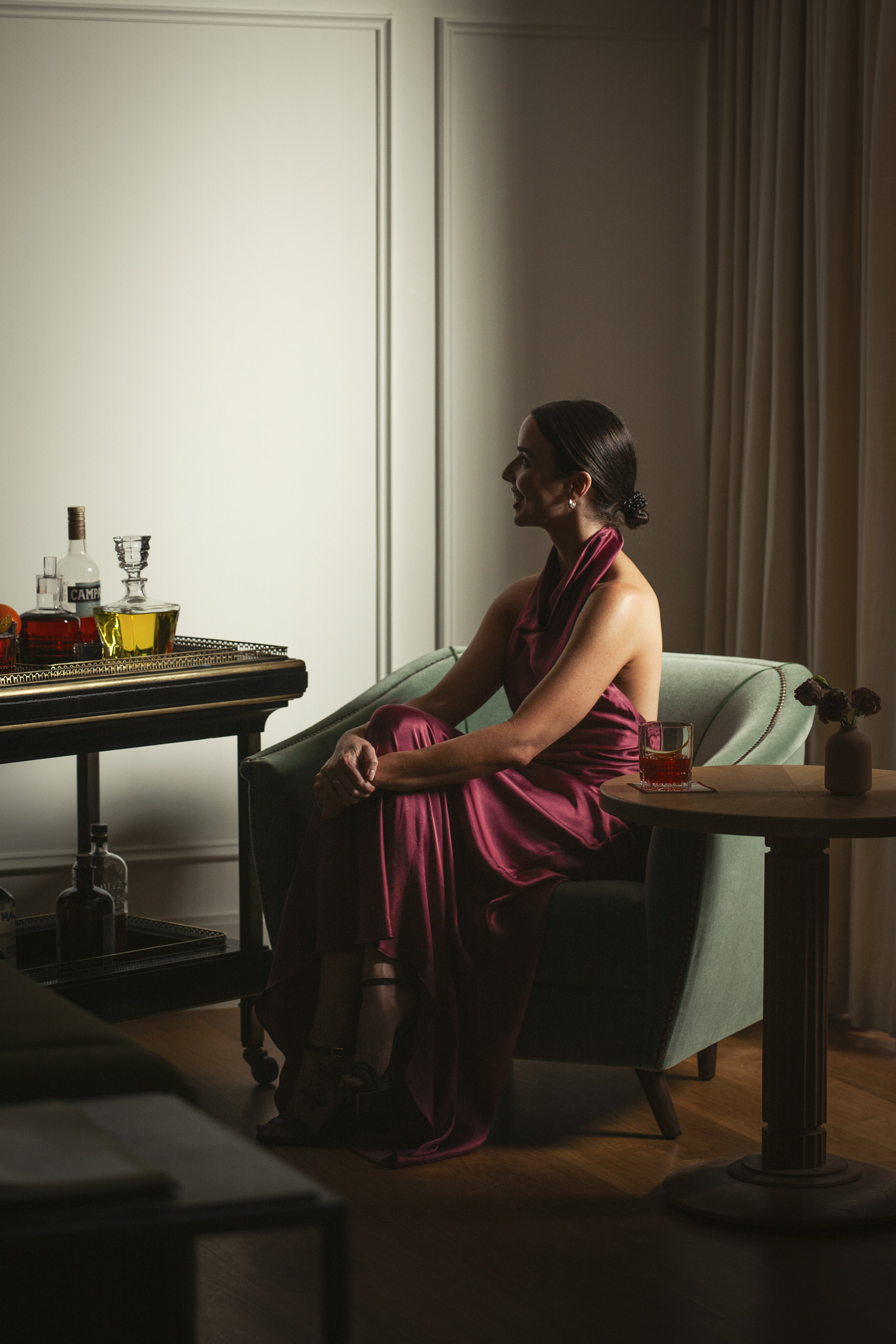Woman seated in armchair beside cocktail cart.