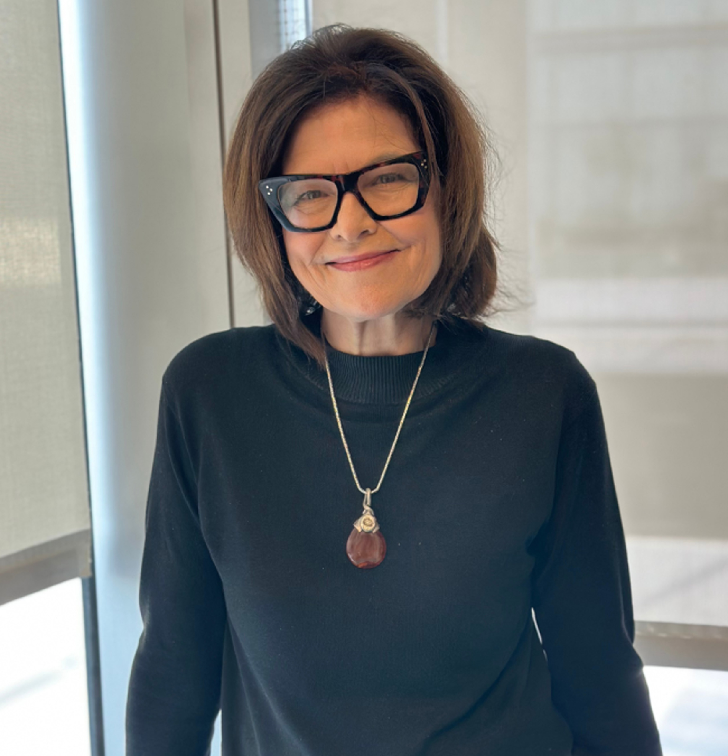 Smiling woman in black sweater and glasses, wearing pendant.