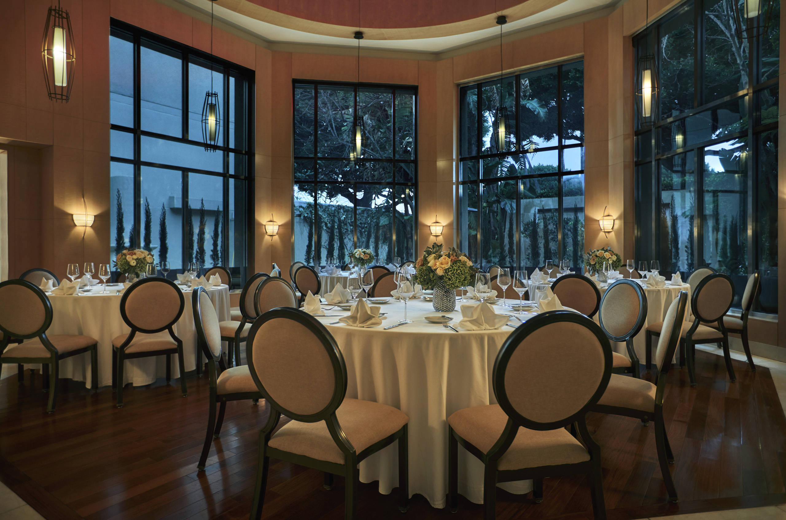 Round banquet tables set for dinner in glass-walled room.