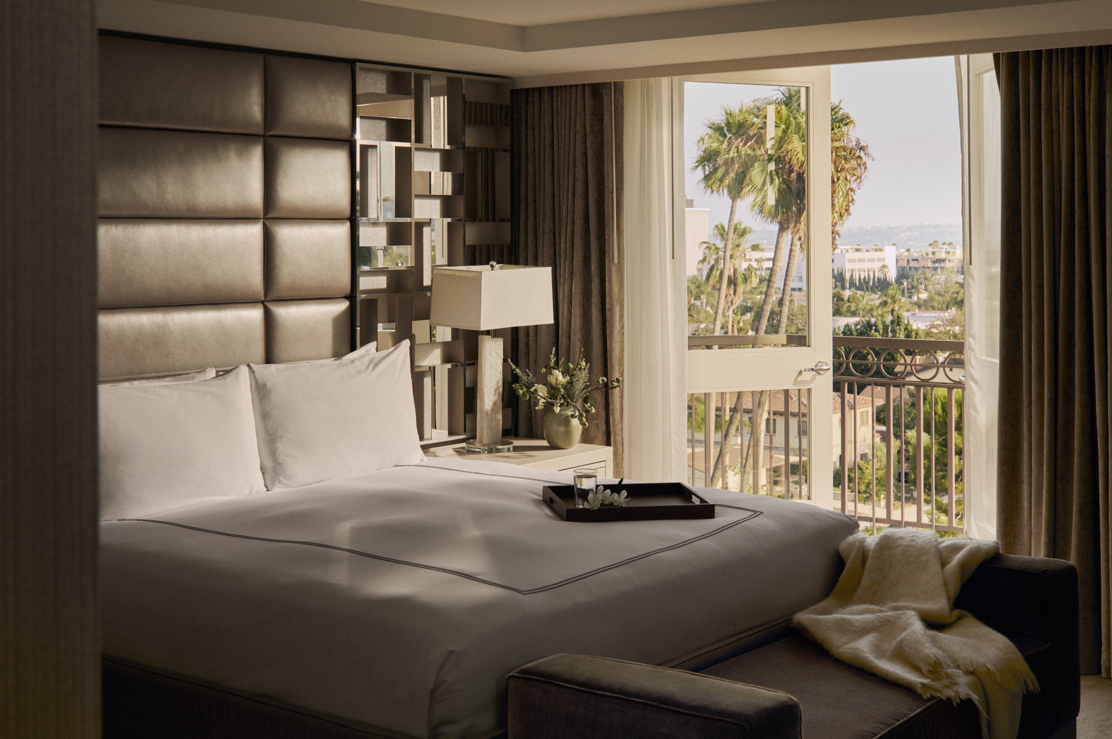 a white bed in a naturally sunlit hotel room with palm trees outside the window